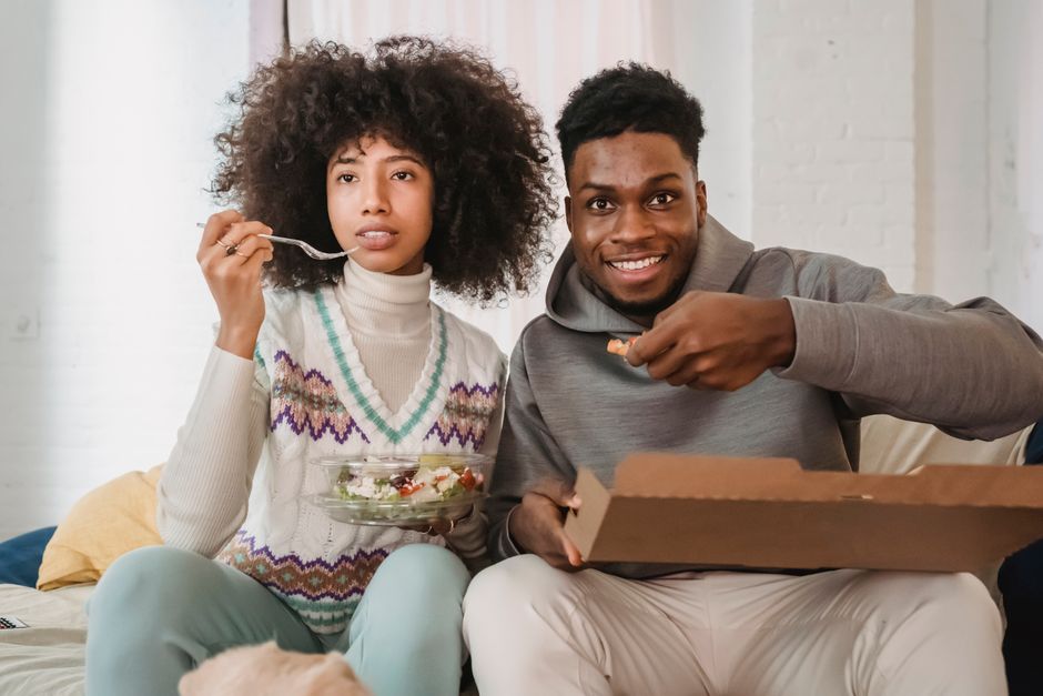 Content African American couple eating salad and pizza in carton box while having fun at home