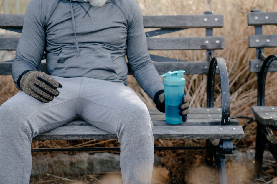 Crop anonymous male in activewear and gloves resting on wooden bench in city park with bottle of water in daylight