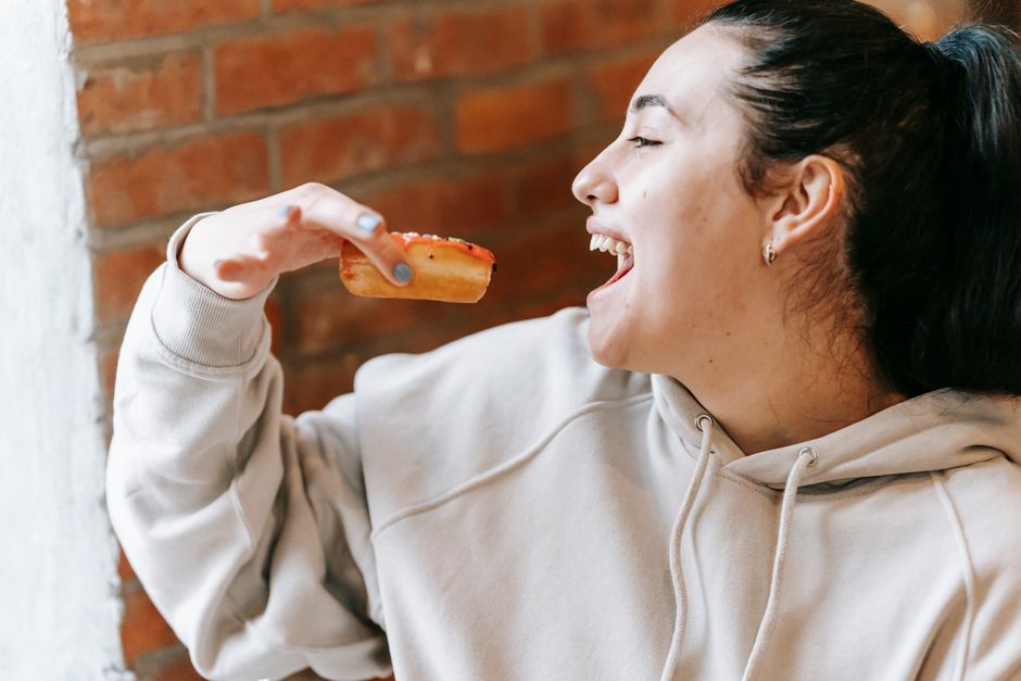 Crop joyful female wearing loose hoodie biting tasty fresh donut in light room