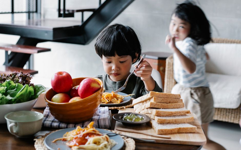 Cute ethnic child eating delicious spaghetti sitting at table with bread and bow of ripe fruits while active sister running around table