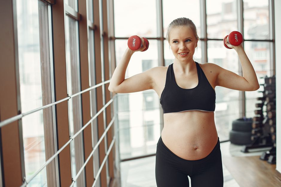 Expectant woman in workout gear lifts dumbbells indoors, embodying fitness and health.