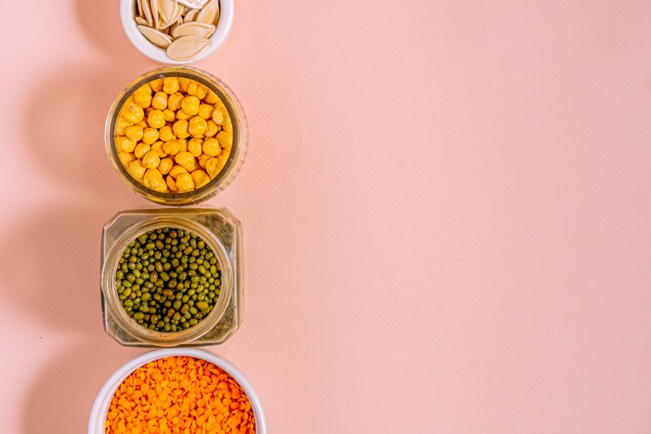Flat lay of chickpeas, lentils, mung beans, and pumpkin seeds with pink backdrop.