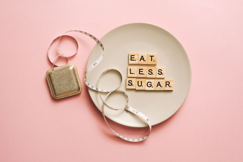 Flat lay of Scrabble tiles saying 'Eat Less Sugar' on a plate with a tape measure on a pink background.