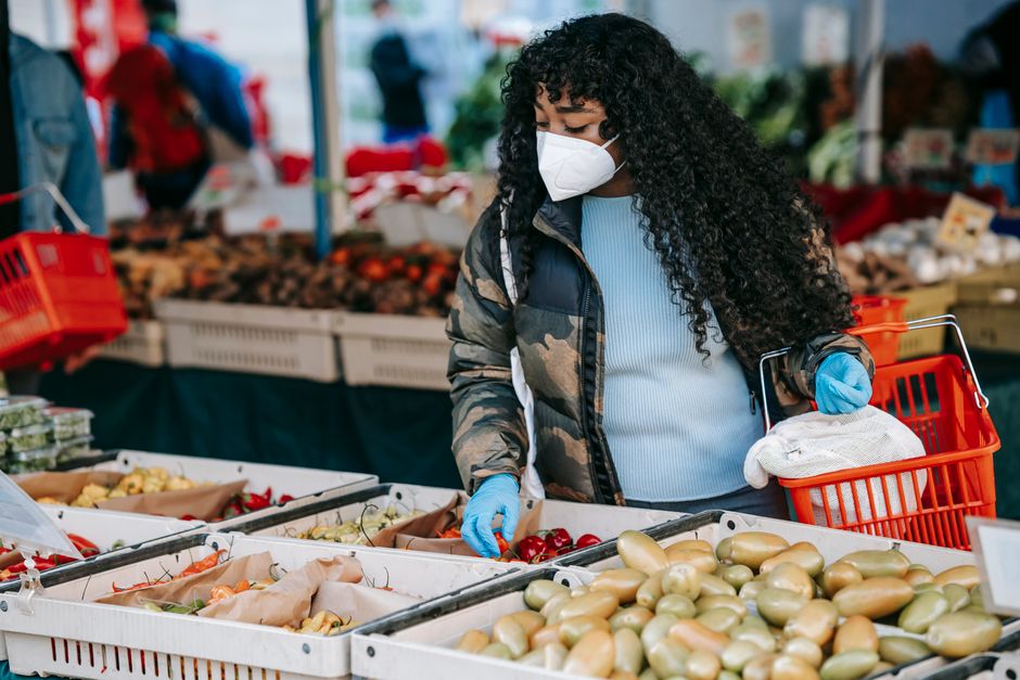 Focused African American female in warm jacket and face mask picking fresh vegetables while doing grocery shopping in supermarket