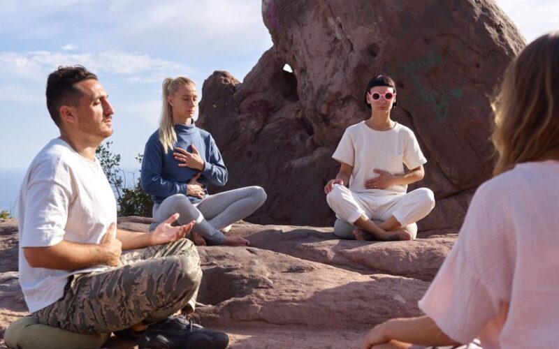 Four people meditate on rocky terrain under a clear sky, promoting serenity and mindfulness.