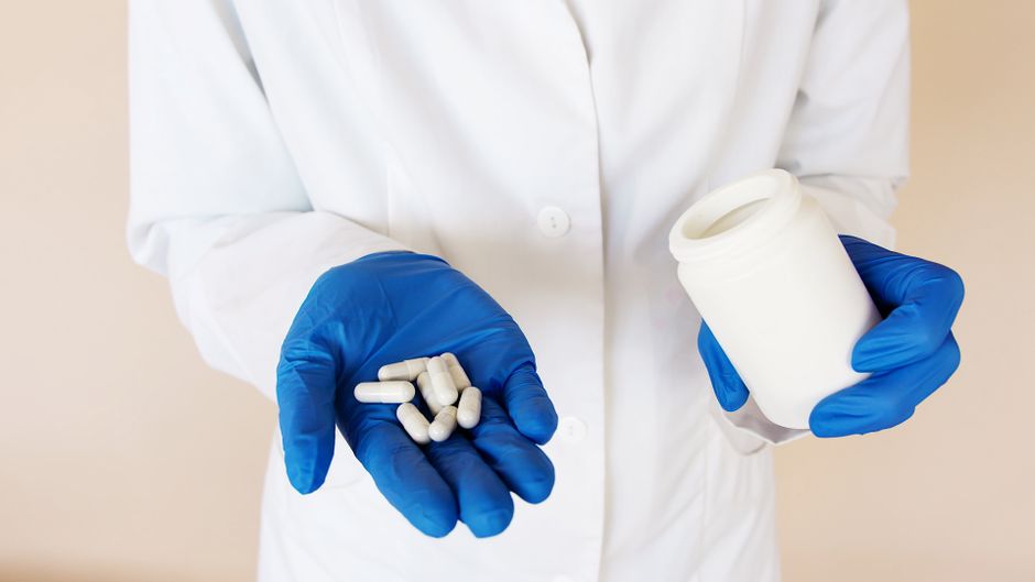 From above crop faceless medical specialist in white coat and blue latex gloves with open plastic bottle in hand and pile of white pills on palm