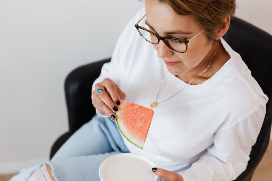 From above of concentrated female in white sweater and eyeglasses sitting in comfortable leather armchair with piece of fresh juicy watermelon