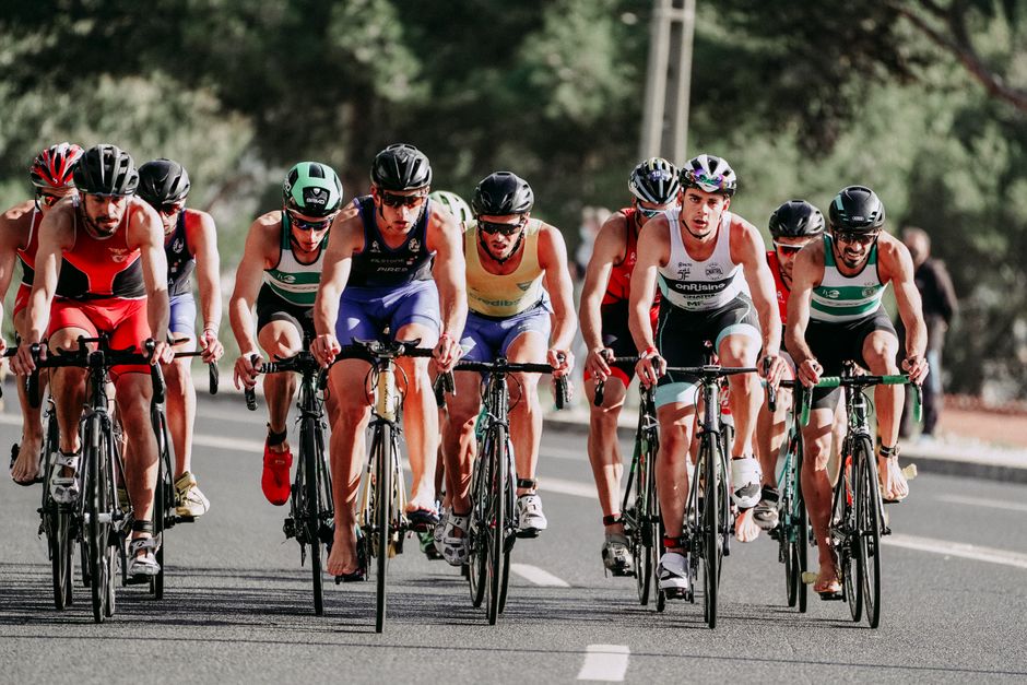 Full body of sportsmen in t shirts and shorts with helmets on heads riding bicycles on even paved road in summertime on blurred background