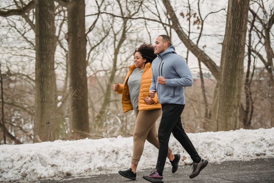Full body side view of plump African American female and black personal coach jogging in winter park with trees during workout