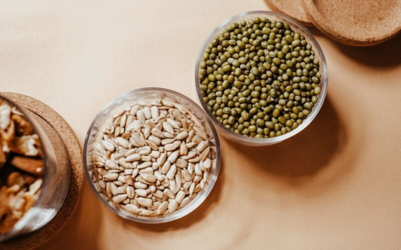 Glass bowls filled with organic mung beans and sunflower seeds on a neutral background.
