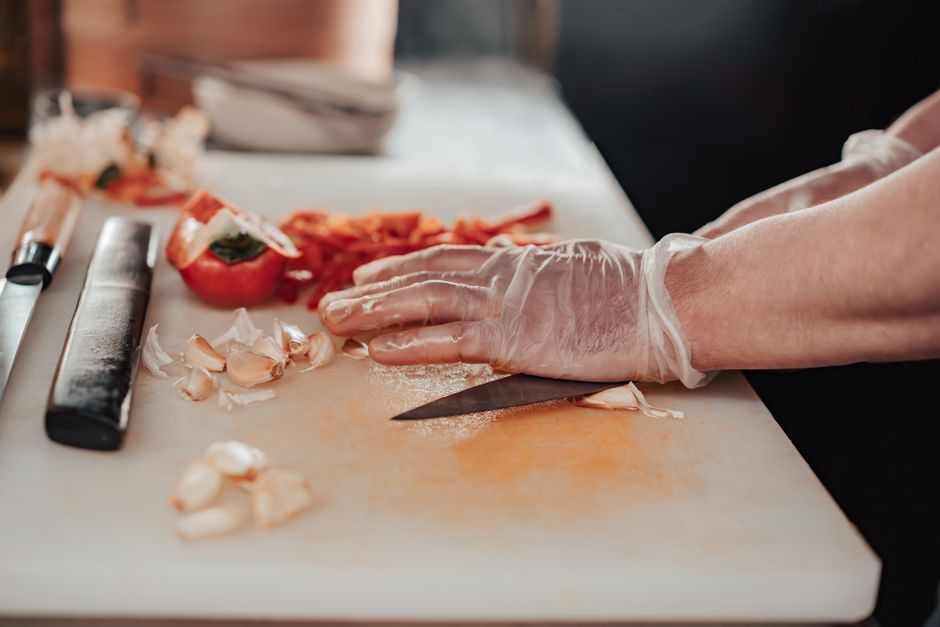 Gloved hands chopping garlic and tomatoes on a cutting board in a kitchen.