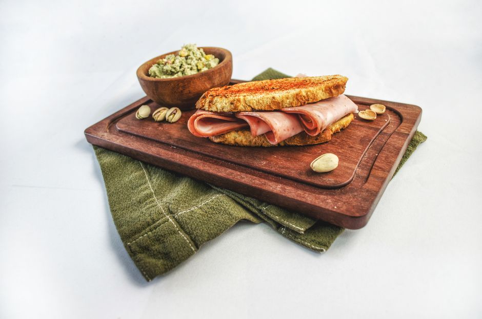 Gourmet ham sandwich with pistachios and dip on wooden board, perfect for food photography.