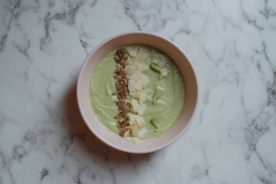 Healthy green smoothie bowl topped with almonds and seeds on marble surface.