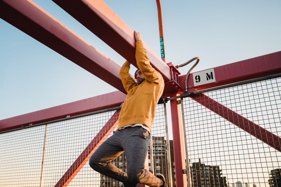 Low angle of young bearded male athlete hanging on beam of city bridge while practicing parkour and looking away