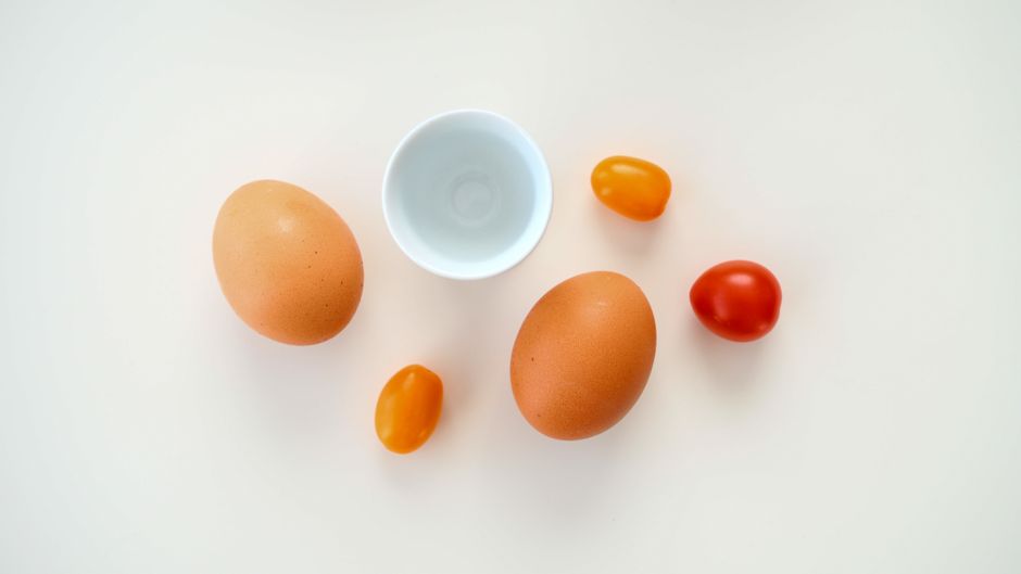 Minimalist flat lay of eggs and cherry tomatoes with a bowl on a white backdrop.
