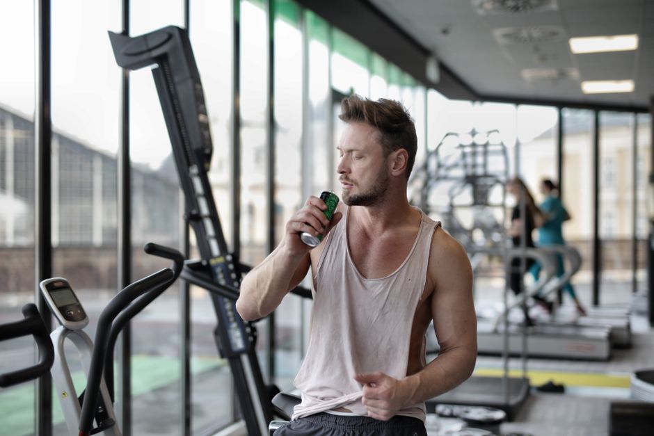 Muscular young sportsman in casual sportswear drinking fresh energy beverage near window while resting after intense workout in modern fitness club