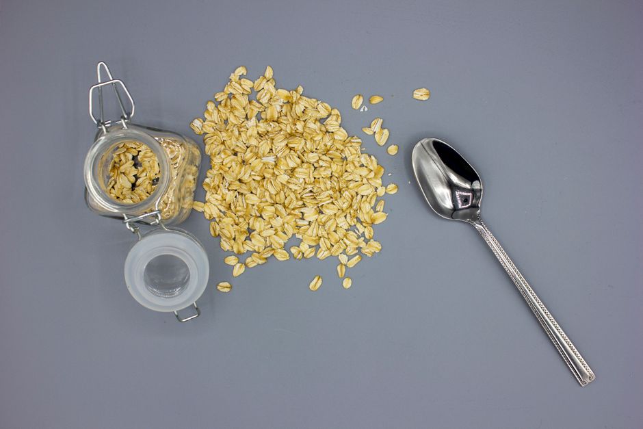 Overhead shot of scattered oats with a glass jar and silver spoon on a gray surface, perfect for breakfast themes.