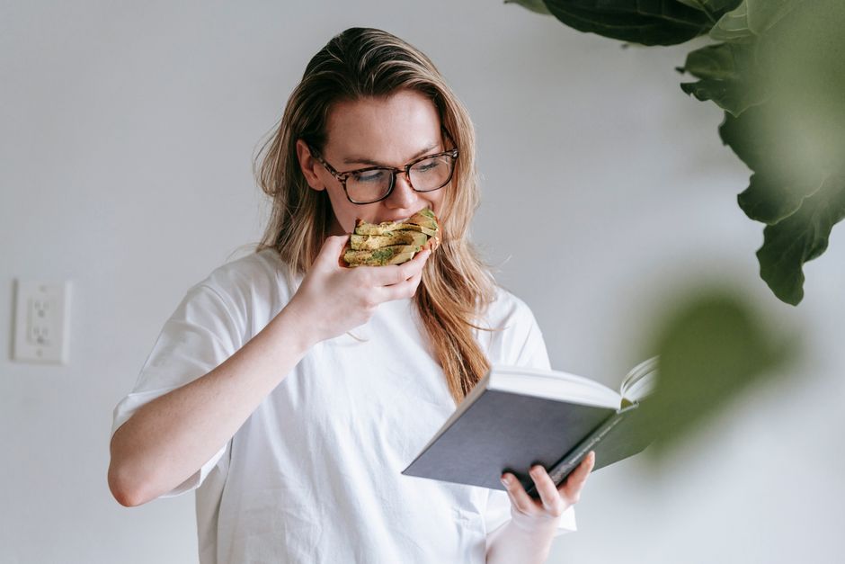 Passionate young female in home white t shirt and eyeglasses immersed in book reading while eating breakfast toast