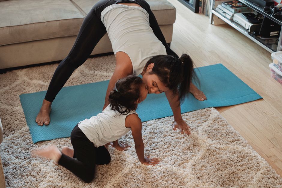 Positive fit Asian female in activewear performing Downward Facing Dog pose and kissing adorable daughter while practicing yoga in living room