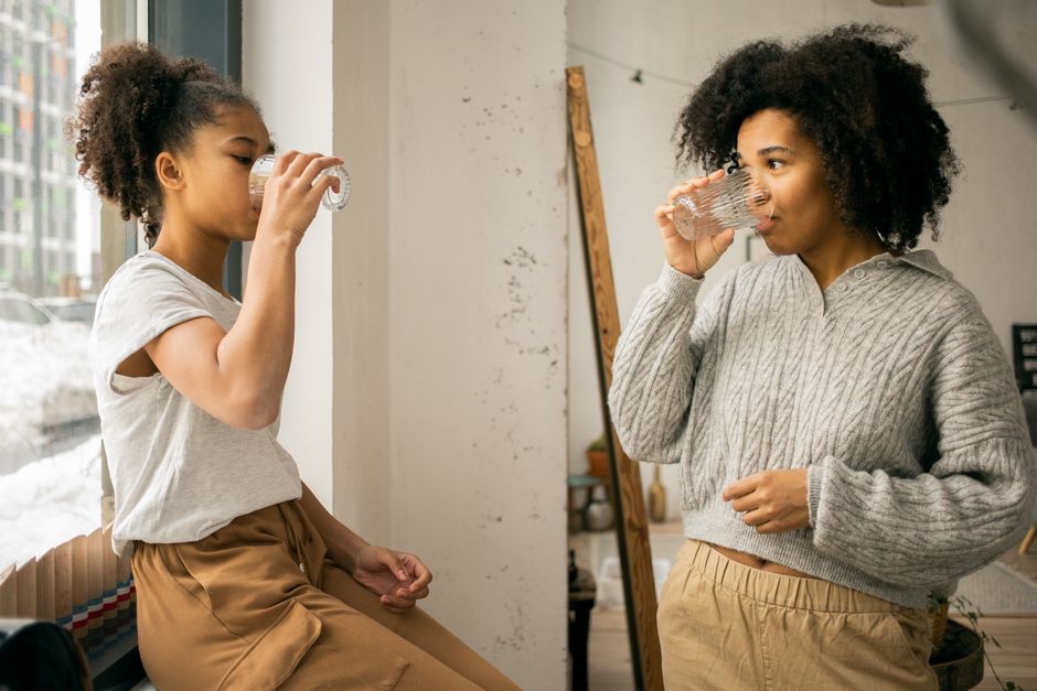 Side view of African American daughter and mother in casual wear drinking water from glasses near window in light room