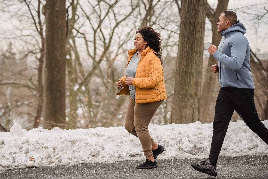 Side view of plump African American female in warm clothes running near black coach on road among tree trunks in winter time