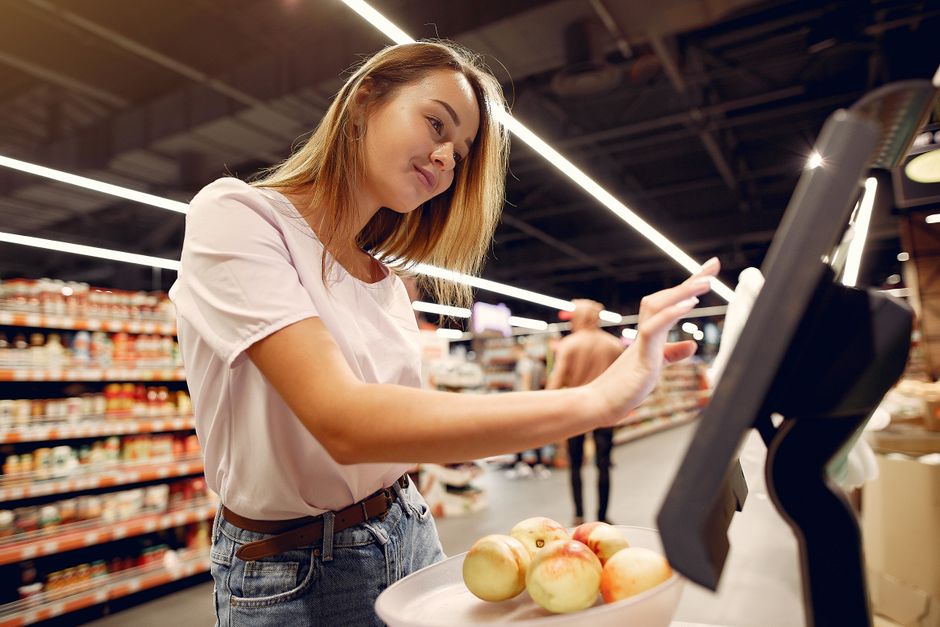 Side view of young woman in trendy clothes weighing peaches on scales while  shopping in supermarket during purchase food