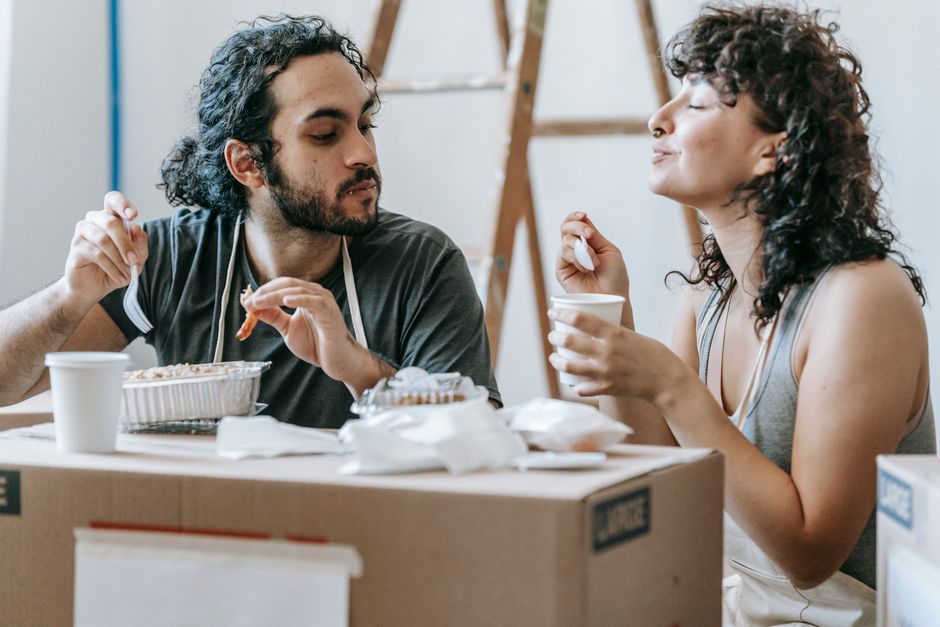 Smiling ethnic woman with closed eyes and coffee against boyfriend with tasty food to go on cardboard box at home