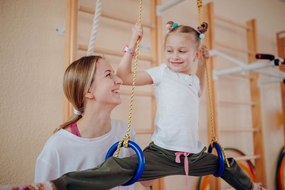 Smiling mother teaches her daughter gymnastics on rings indoors, fostering bonding and flexibility.
