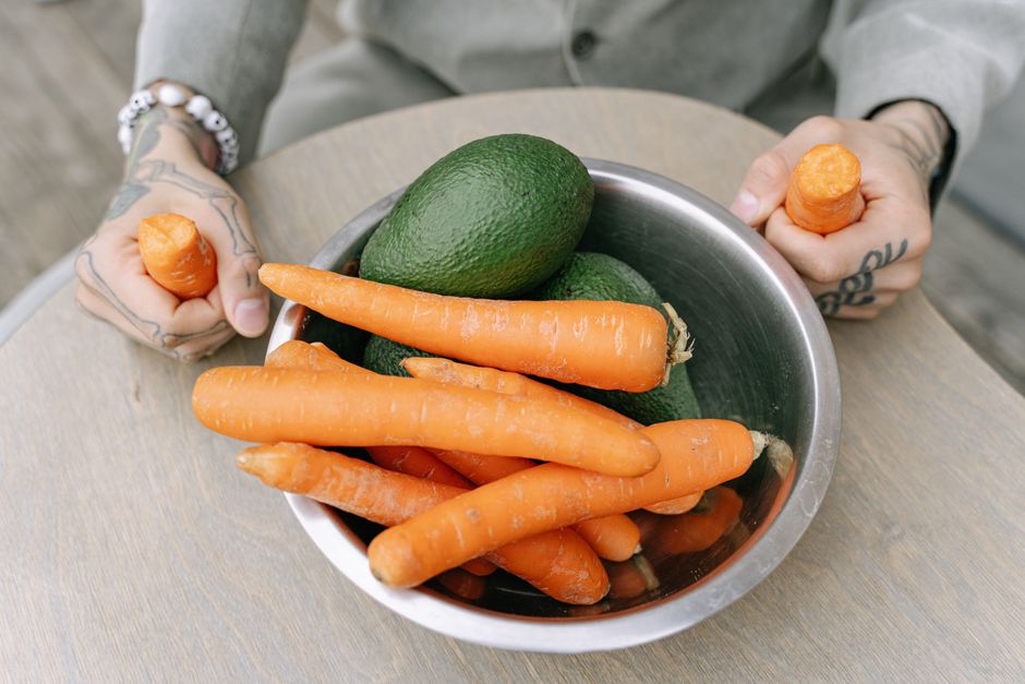 Tattooed person holding a steel bowl filled with fresh avocados and carrots on a wooden table.