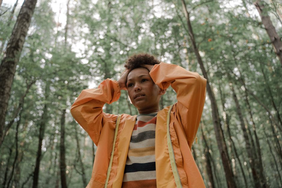 Teen holding head in thinking pose, wearing yellow raincoat, surrounded by forest greenery.