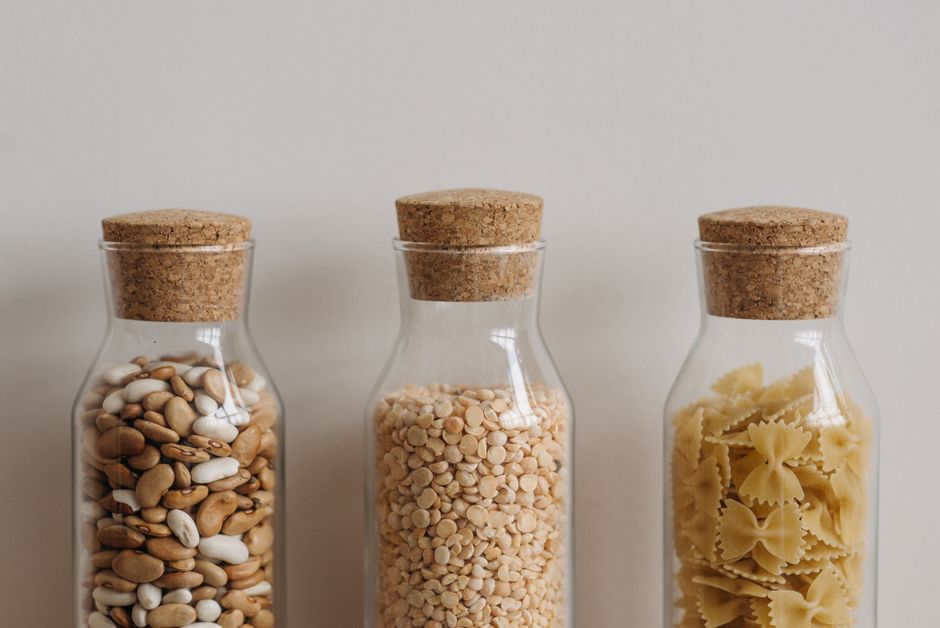 Three clear glass jars with cork lids hold beans, lentils, and pasta showcasing pantry organization.