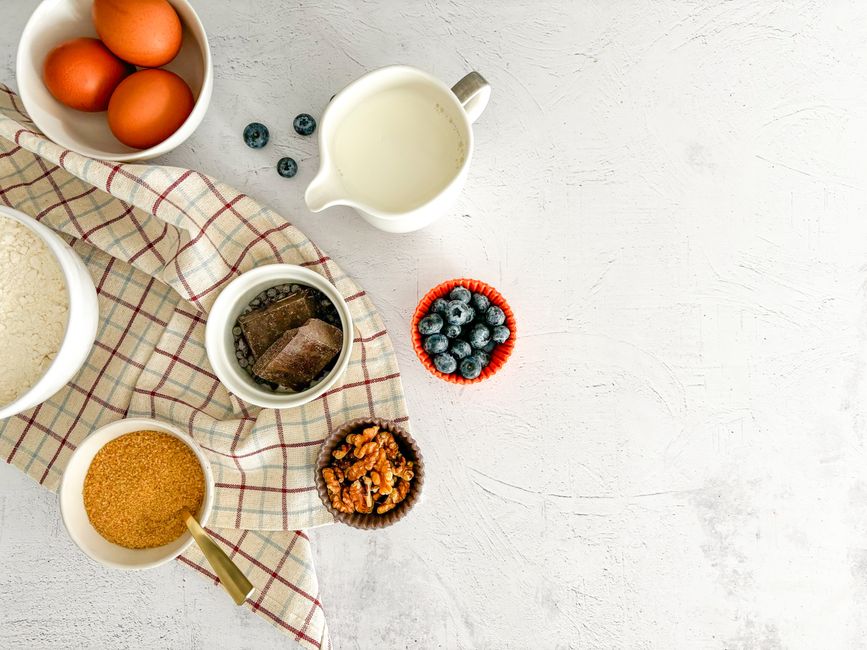 Top view of baking ingredients including blueberries, chocolate, eggs, and milk on a white surface.