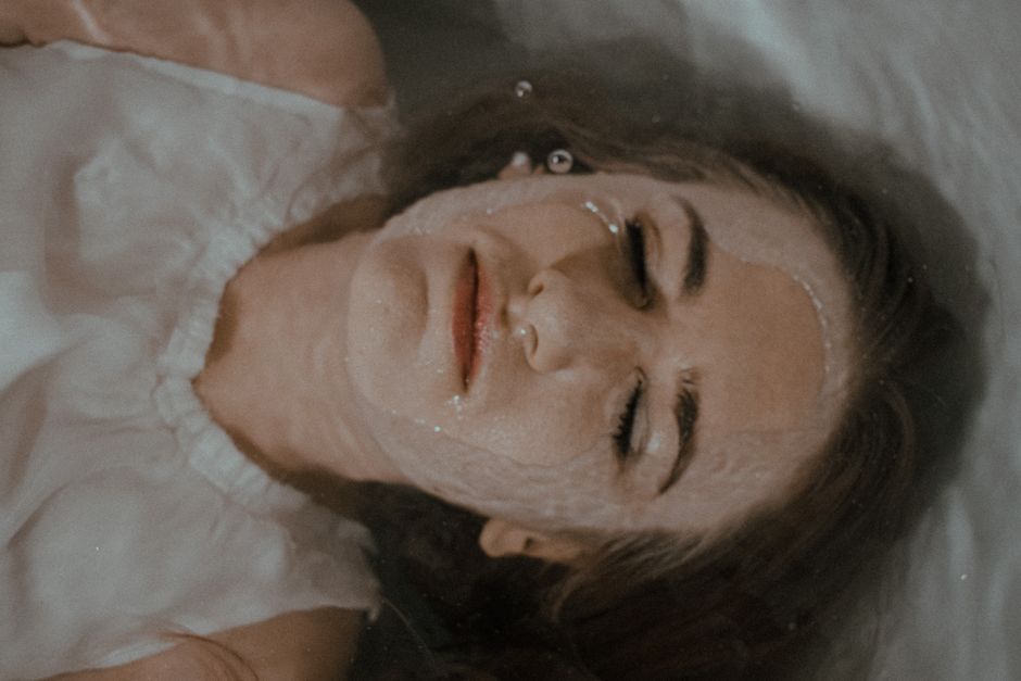 Top view of female wearing white outfit with closed eyes lying in bathtub with head immersed in water in bathroom