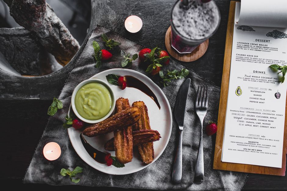 Top view of fried churro and avocado sauce on plate garnished with chocolate and strawberry and mint leaves served with lavender dessert in glass on table with knife and fork and menu and hole for plant trunk and candles