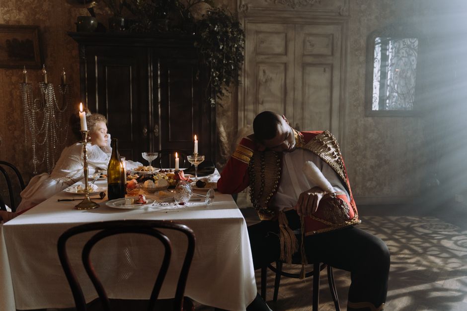 Two adults in period costumes asleep after a party at an elegant dining room table.