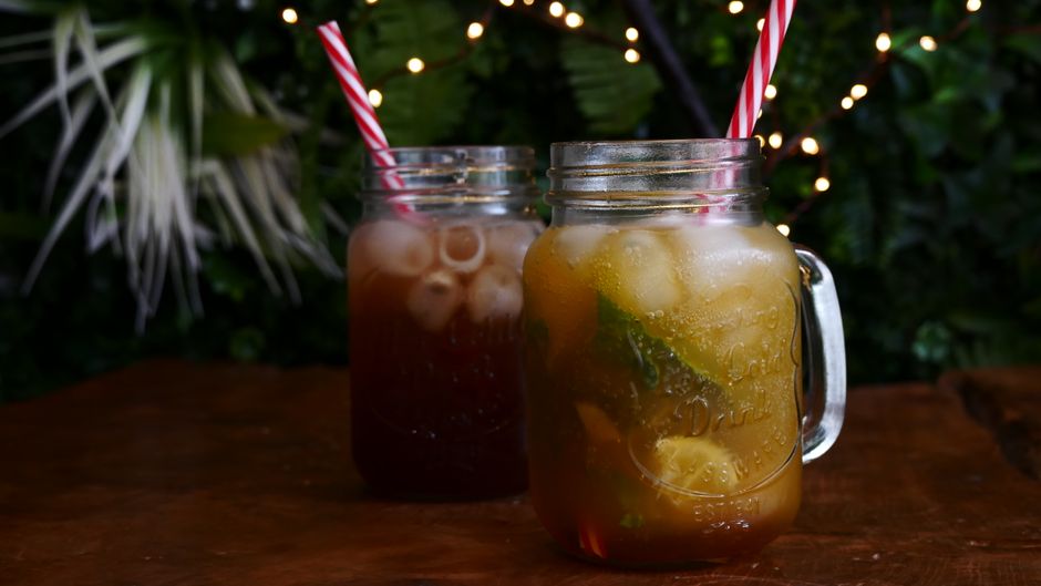 Two iced beverages with straws in mason jars on a wooden table outdoors.