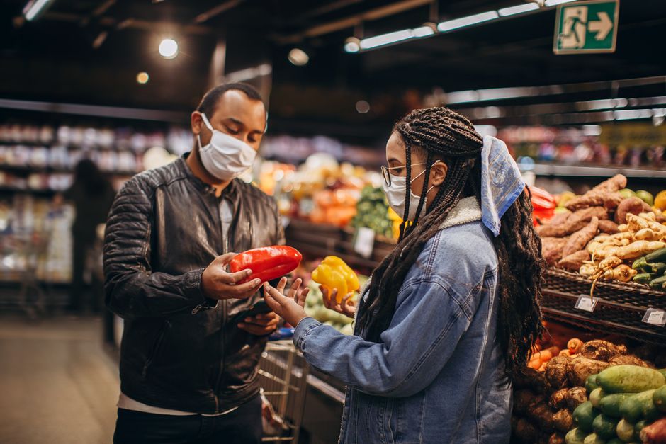 Two masked individuals selecting fresh peppers in a supermarket aisle, practicing safety during pandemic.