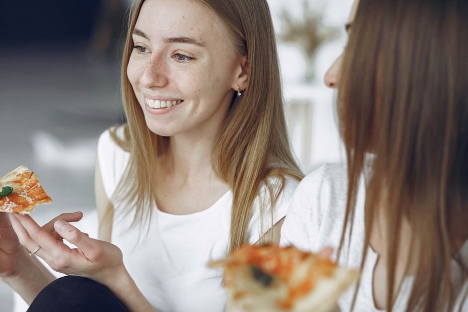 Two young women smiling and enjoying pizza together indoors. A moment of joy and friendship.