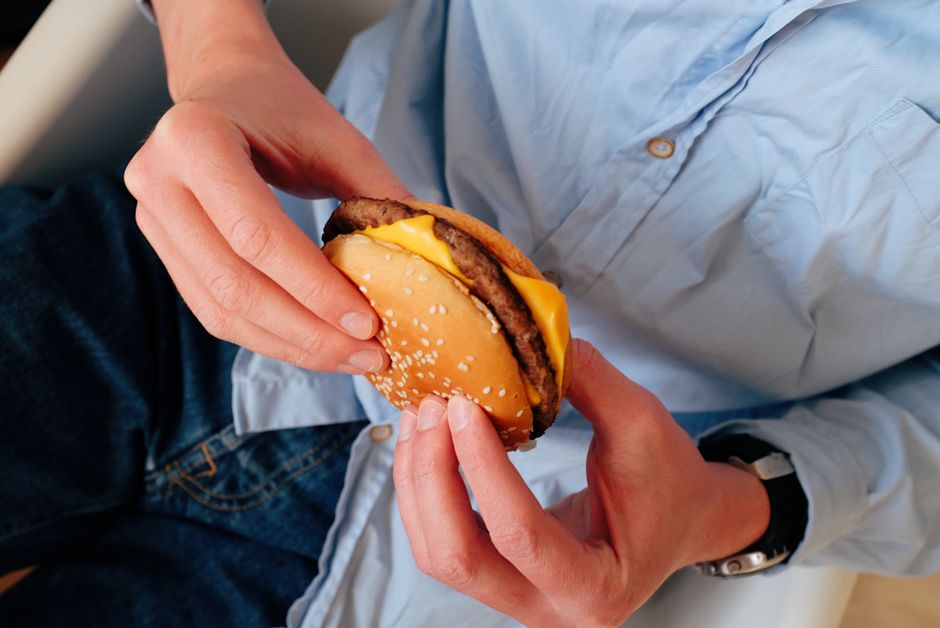 Unrecognizable male in blue shirt sitting on chair and holding delicious burger with cheese and cutlet in hands