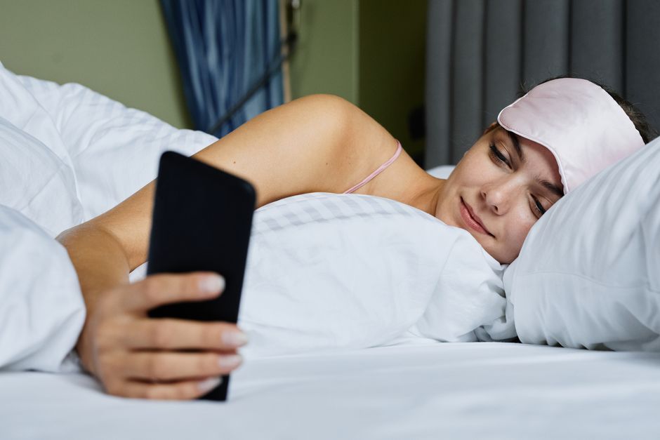 Woman in bed wearing an eye mask and using a smartphone, conveying relaxation.