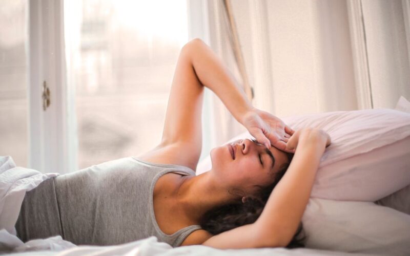 Woman in gray tank top resting on bed with natural light from window, looking relaxed and comfortable.