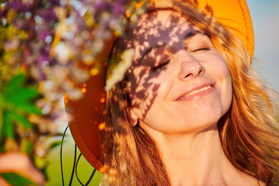 Woman in sun hat enjoying sunlight, surrounded by nature, with shadows on her face.