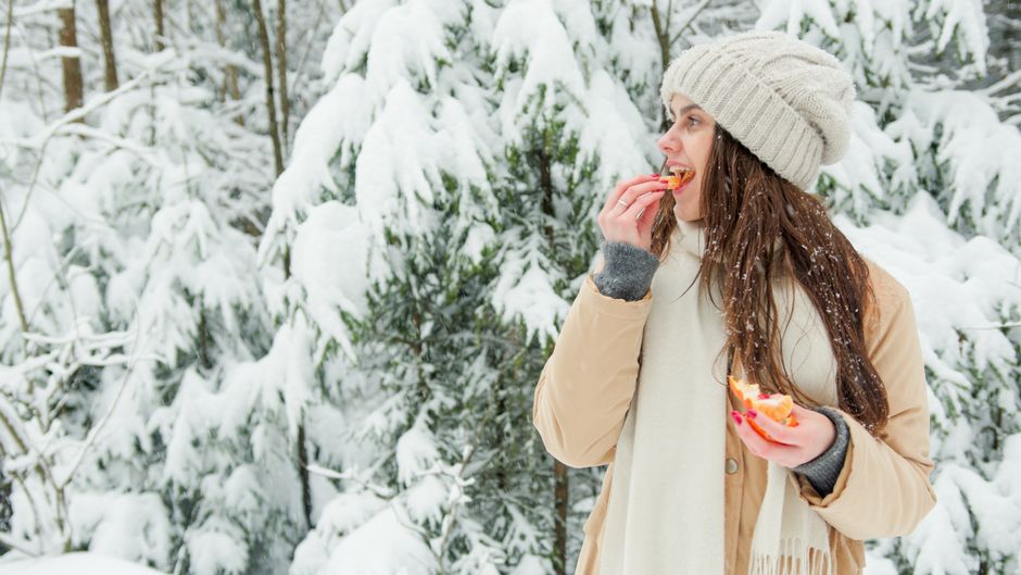 Woman in winter clothing eating an orange in a snow-covered forest.