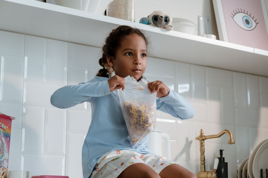 Young girl sitting in kitchen holding cereal bag, ready for breakfast.