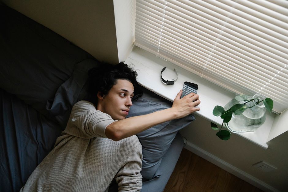 Young man lying on bed using smartphone with blinds open, showcasing modern indoor lifestyle.