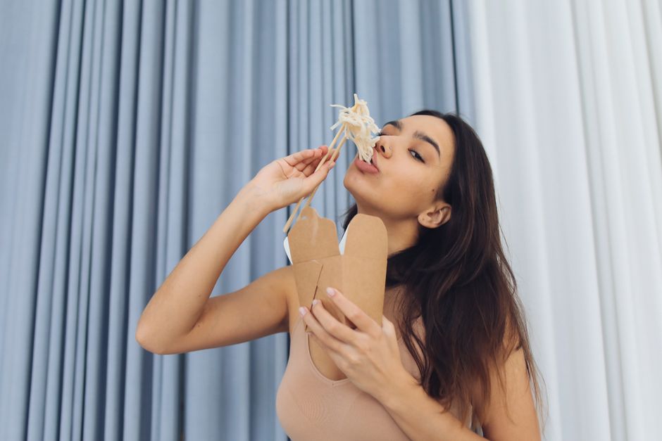 Young woman savoring noodles with chopsticks, enjoying a casual indoor meal in front of a curtain background.