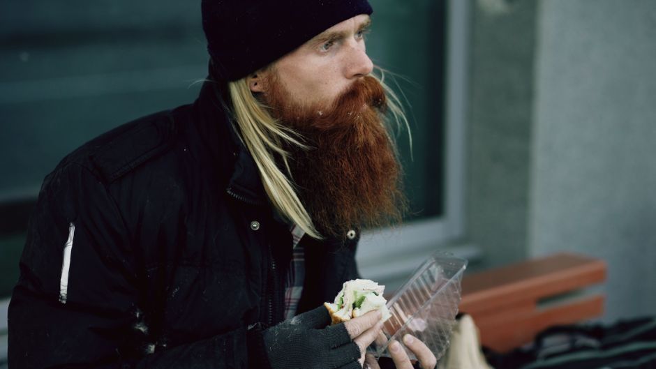 A bearded man enjoys a sandwich while seated outdoors, dressed warmly for winter.