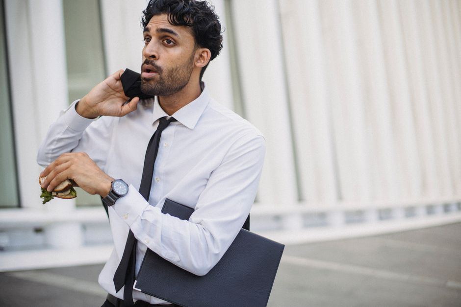 A businessman in corporate attire multitasking with a sandwich and phone outdoors.