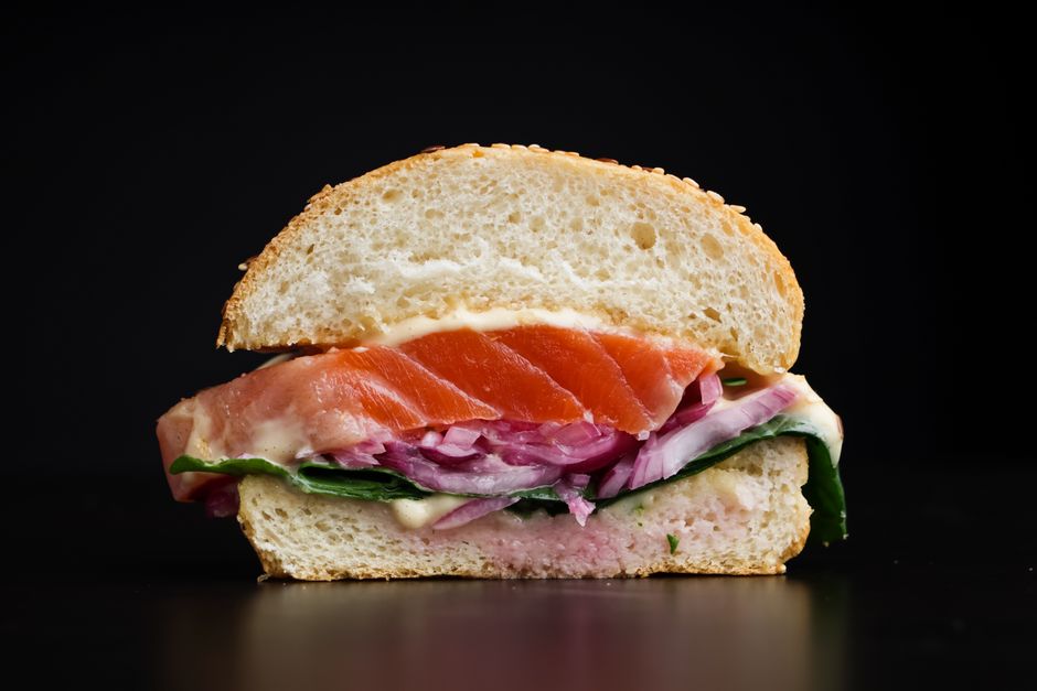A close-up of a gourmet sandwich with salmon, onions, and greens on a black backdrop.