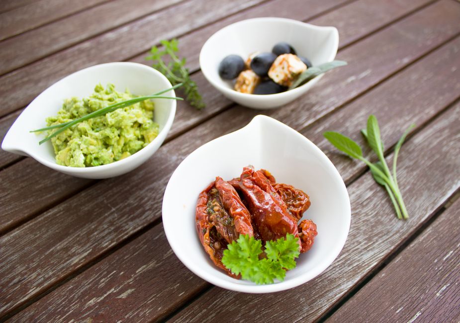 A close-up of three flavorful bowls on a wooden table featuring fresh guacamole, sundried tomatoes, and olives with cheese.